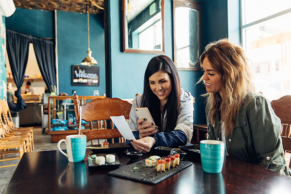 Two women of the Millennial Generation are taking a picture of a restaurant receipt after eating lunch at a local sushi restaurant. The woman is using her bank's app to balance her monthly budget.