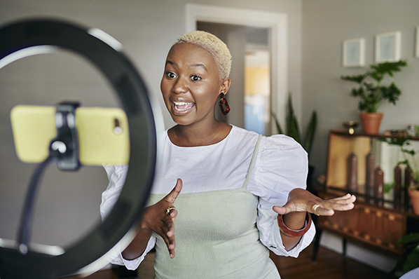 Young lady doing a dance in her living room while smiling in front of a cell phone docked in a ring light.