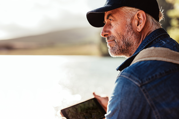 Older male holding a hand-held electronic while looking thoughtfully over a lake.