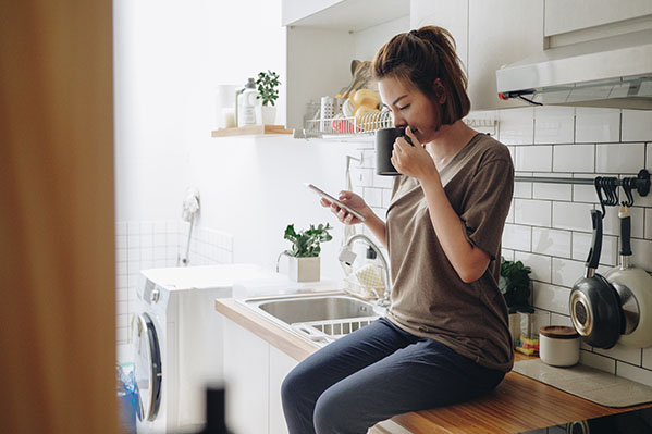Woman sitting in a cozy kitchen while drinking coffee and looking at her cell phone.