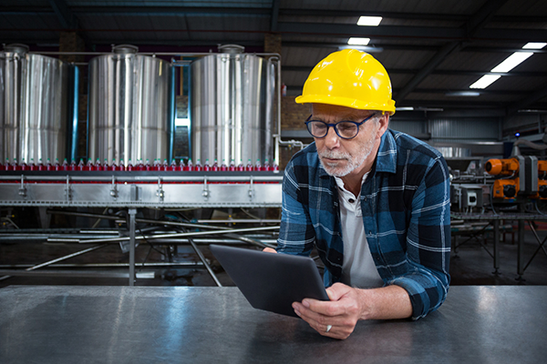 middle-aged plant worker in a plaid shirt and a safety helmet leaning on the counter with a tablet in hand