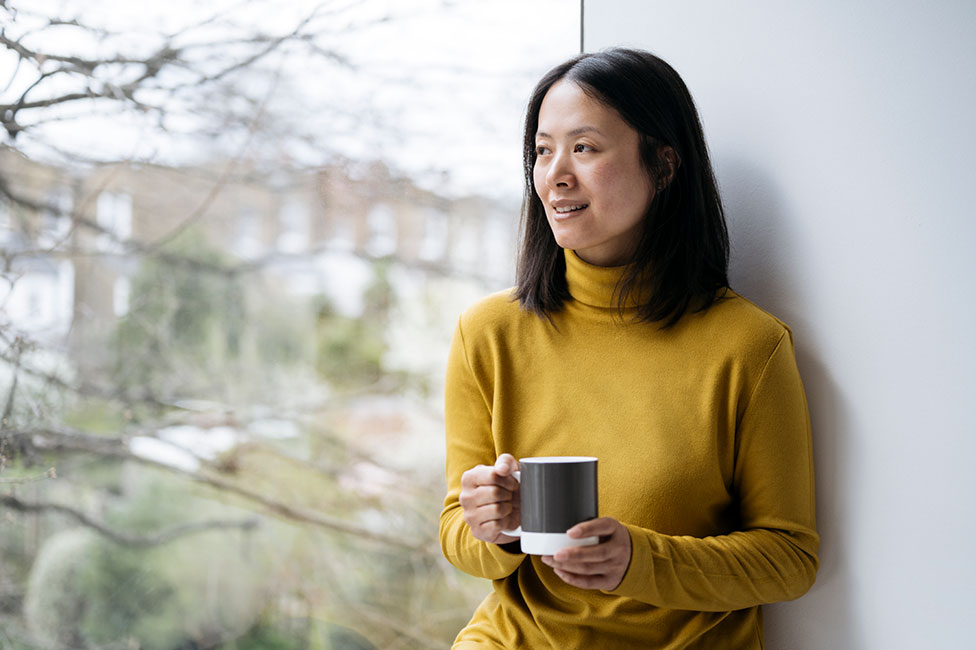 middle aged asian woman in a yellow sweater holding a mug and looking out the window
